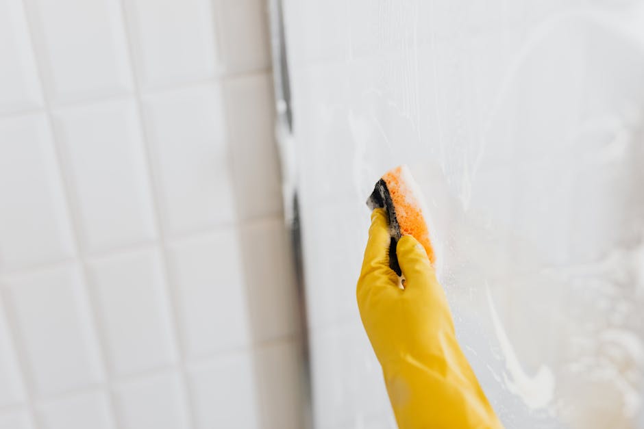 From above of crop anonymous person in yellow rubber protective glove washing shower cabin glass with sponge and detergent